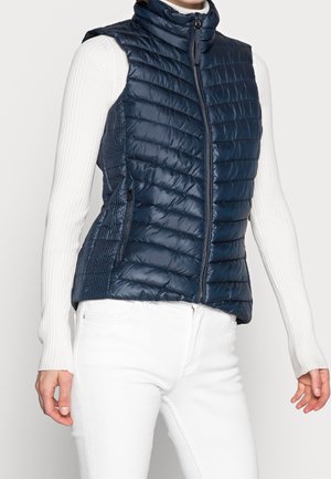 Femme portant une veste matelassée sans manches bleu marine sur un haut blanc à manches longues côtelé et un pantalon blanc, debout devant un fond uni.