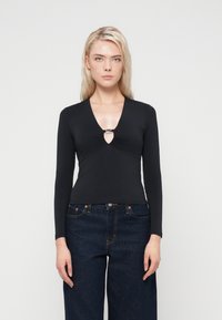 Haut noir à manches longues avec un décolleté en V profond, doté d'un détail en métal argenté et d'un tissu froncé à l'avant, porté avec un pantalon en denim foncé.