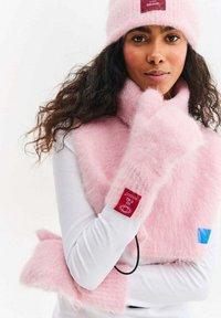 Różowa puszysta czapka i pasujący szalik z metką; biała bluzka z długim rękawem. Szalik owija się wokół szyi, a rękawiczki wykonane są z tego samego materiału i mają ten sam wzór.