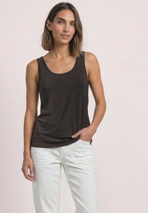 Mujer con pelo castaño hasta los hombros, que lleva una camiseta negra sin mangas y pantalones blancos, de pie con una mano en el bolsillo.