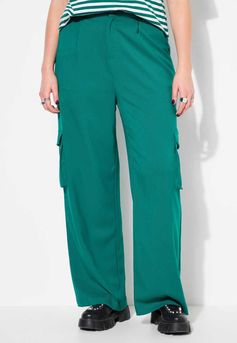 Breite grüne Hose aus einem glatten Stoff, mit seitlichen Cargotaschen und einem lässigen Schnitt. Kombiniert mit schwarzen klobigen Schuhen.