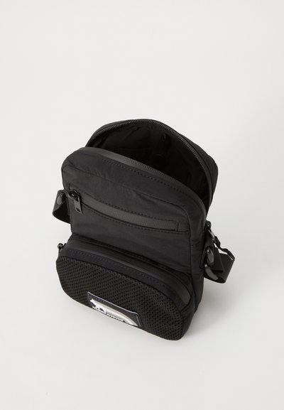 Sac en nylon noir avec un compartiment principal zippé, une poche avant en maille et une bandoulière réglable. Caractéristiques d'un design compact et rectangulaire.