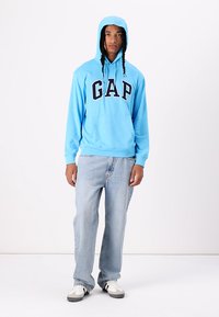 Blue hoodie featuring "GAP" logo in black, with a kangaroo pocket. Pair of light blue jeans and white sneakers with black accents.