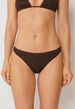 Frau trägt ein dunkelbraunes, glitzerndes Bikini-Set, zeigt Oberkörper und Oberschenkel vor einem schlichten hellen Hintergrund.