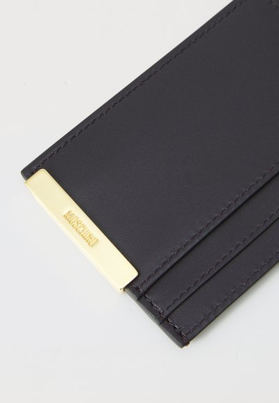 Cardholder din piele de culoare mov închis, având patru sloturi și un accent metalic de ton auriu cu logo embosat. Textură netedă și cusături precise.