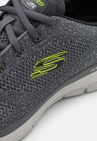 Grauer Sportschuh mit gewobenem Mesh-Obermaterial, grünem Logo-Akzent und grauer Gummisohle mit strukturiertem Profil.