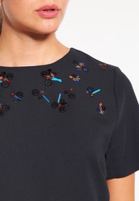 Top blu scuro con applicazioni floreali, paillettes nere, perline colorate e dettagli testurizzati sul collo e sulle spalle.