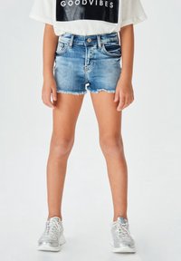 Shorts in denim blu medio con orlo sfrangiato, tasche e chiusura con bottone. Abbinati a scarpe da ginnastica argentate e una maglietta bianca con grafica.