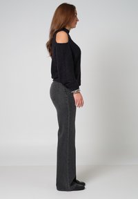 Schwarzer Cold-Shoulder-Pullover aus strukturiertem Stoff, kombiniert mit hochgeschnittenen, dunkelgrauen Schlagjeans und schwarzen flachen Schuhen.