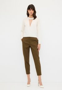 Weißes Langarmoberteil mit tiefem V-Ausschnitt und gestricktem Saum, kombiniert mit figurschmeichelnden olivgrünen Hosen. Die Modell trägt weiße spitz zulaufende High Heels.