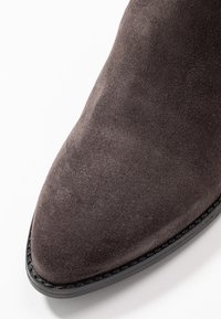 Bottines en daim gris foncé avec un bout pointu, présentant une finition lisse et une semelle en caoutchouc noir avec des coutures décoratives le long du bord.