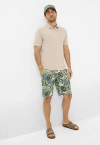 Maglietta polo beige a maniche corte, pantaloni corti verdi e blu con motivo di foglie tropicali, sandali scivolati marroni e un berretto verde.