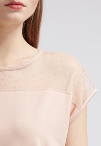 Růžový top v odstínu blush se síťovanou průhlednou overlay s puntíky na ramenou, hladkým tělem z jednolitého materiálu a jednoduchým kulatým výstřihem.