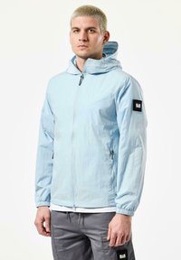 Light blue windbreaker with a zip front, elastic cuffs, and a hood. Features black patch on left sleeve and zippered pockets. Smooth texture.