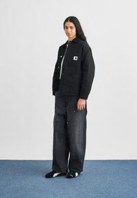 Mladý človek stojí na modrom koberci, má na sebe čiernu bundu Carhartt, voľné tmavo sivé džínsy a čierne tenisky Puma, na pozadí je biela stena.