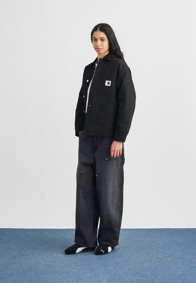 Joven de pie sobre una alfombra azul, vistiendo una chaqueta negra Carhartt, jeans oscuros y holgados, y zapatillas negras Puma, frente a una pared blanca.