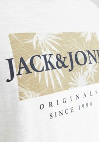 T-shirt blanc à manches courtes avec un graphique beige réhaussé de motifs de feuilles de palmier et du texte noir indiquant "JACK & JONES ORIGINALS SINCE 1990."