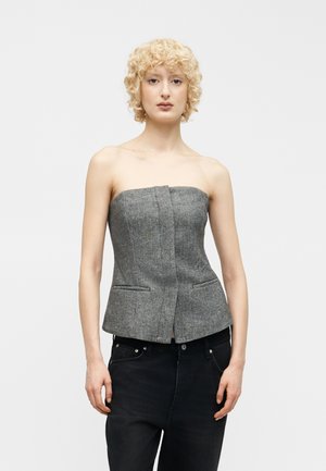 Młoda osoba o krótkich, kręconych blond włosach, ubrana w szary, dopasowany gorset bez ramiączek i czarne dżinsy, stojąca na białym tle.