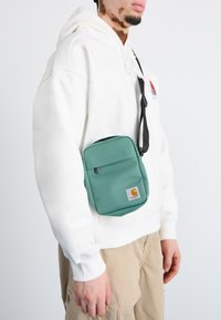 Green fabric crossbody bag with a front zipper pocket, logo patch, and adjustable strap, worn over a white hoodie. Smooth texture.