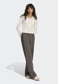 Žena s kučeravými vlasmi, oblečená v bielej blúzke s dlhým rukávom, šedých športových nohaviciach s čiernymi pruhmi a bielych teniskách, stojí so zdvihnutými rukami za chrbtom.