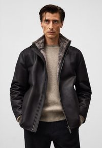 Mężczyzna w czarnej skórzanej kurtce z futrzanym wykończeniem, noszący beżowy sweter, stoi z rękami w kieszeniach na jasnym, jednolitym tle.