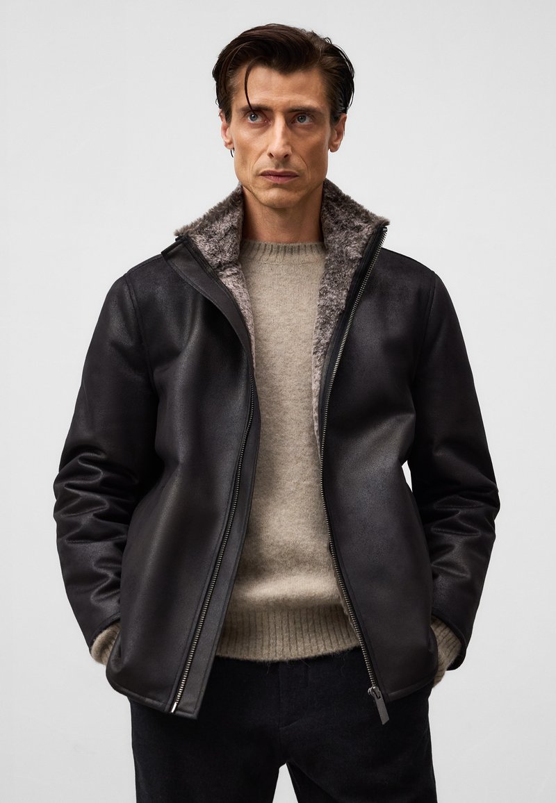 Mężczyzna w czarnej skórzanej kurtce z futrzanym wykończeniem, noszący beżowy sweter, stoi z rękami w kieszeniach na jasnym, jednolitym tle.