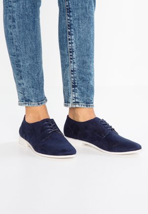 Person iført blå ruskindssnøresko og opsmøgede blå jeans står på en hvid overflade.