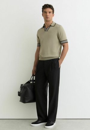 Man staat tegen een witte muur, draagt een beige gebreide polo met korte mouwen, zwarte broek, zwarte sneakers en houdt een grote zwarte leren reistas vast.