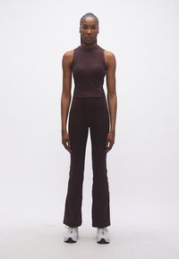 Γυναίκα που στέκεται με το πρόσωπο προς τα εμπρός, φορώντας καφέ αμάνικη μπλούζα και καφέ παντελόνι με φουσκωτή εφαρμογή, με λευκά αθλητικά παπούτσια, σε λευκό φόντο.