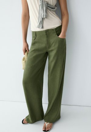 Persona che indossa pantaloni verde oliva a vita alta e gamba larga, top beige con maglione grigio sulle spalle, tiene una borsa intrecciata e indossa infradito bianchi.