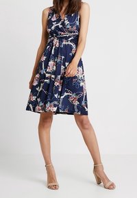 Marineblaues Blumenkleid mit Wickel-Oberteil, V-Ausschnitt und schwingendem Saum. Mit rosa und weißen Blumendrucken. Kurz geschnitten.