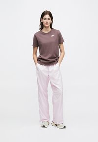 Γυναίκα που στέκεται με τα �χέρια της στις τσέπες, φορώντας ένα καφέ μπλουζάκι Nike, ανοιχτό ροζ παντελόνι και μπεζ αθλητικά παπούτσια σε λευκό φόντο.