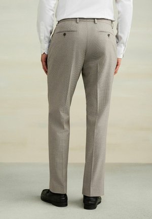 Férfi bézs kockás öltönynadrágban, fehér ingben és fekete elegáns cipőben, háttal áll egy egyszerű világos háttér előtt.