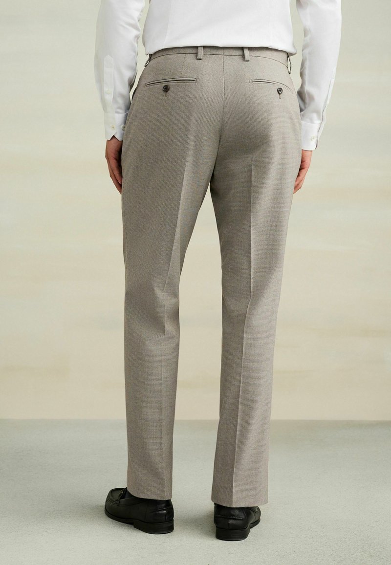 Homme portant un pantalon de costume beige à carreaux, une chemise blanche et des chaussures habillées noires, debout de dos devant un fond clair uni.