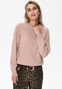 Kobieta z blond włosami, ubrana w sweter z różowego, lekko zakurzonego dzianiny z wzorzystymi otworami oraz spodnie w panterkę z brązowym paskiem.