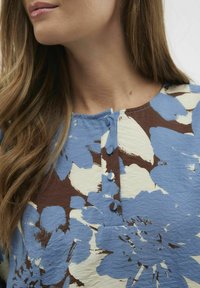 Blumenbluse in Blau, Braun und Creme. Sie hat einen runden Halsausschnitt und drei Knöpfe vorne. Glattes, strukturiertes Material.