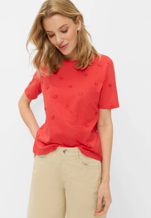 Vrouw met blond haar die een rood T-shirt met korte mouwen draagt met kleine geborduurde bloemen en beige broek tegen een witte achtergrond.