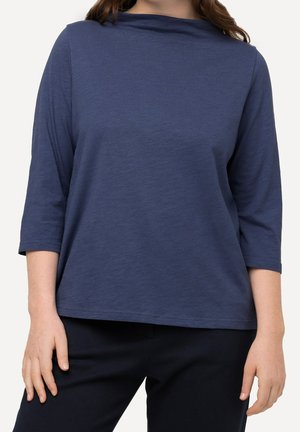 Femme portant un t-shirt bleu uni à manches longues avec un col bateau et un pantalon noir, debout, les mains détendues le long du corps.