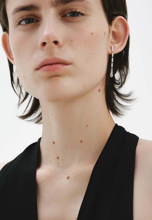 Persona con cabello corto y oscuro que lleva una blusa negra y un pendiente largo plateado, con lunares visibles de piel clara en el cuello y el rostro.