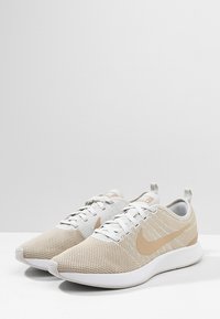 Beiga och vita träningsskor gjorda av ventilerande mesh med en slät sula, med ett framträdande tan Nike-swoosh och snörning.
