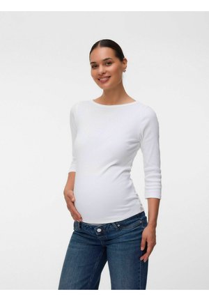 Femme enceinte souriante, portant une chemise blanche à manches longues et un jean bleu, une main reposant sur son ventre sur un fond uni.