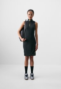 Rochie neagră fără mâneci, cu guler înalt și detalii cu fermoar aurie, având un design tip enwrap și un model subtil lângă tiv. Modelul ține o rucsac negru.
