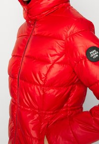 Röd puffjacka med hög krage, dragkedjestängning och elastiska muddar. Har en svart cirkulär logotyp på vänster ärm. Slät, glänsande yta.