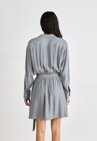 Robe grise à manches longues avec une taille cintrée, des poignets boutonnés et une coupe décontractée. Fabriquée en tissu doux avec un léger brillant et un motif texturé.