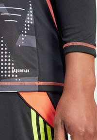 Maglietta sportiva nera con motivi geometrici in grigio e bianco, dettagli arancioni e branding AEROREADY vicino all'orlo. Tessuto testurizzato.