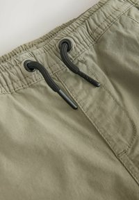 Pantaloni shorts verde oliva in un tessuto testurizzato, con lacci neri e occhielli in vita, progettati per comfort e regolabilità.