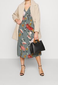 Mujer con abrigo de felpa beige, vestido midi floral verde, sandalias de tacón negras, sosteniendo un bolso negro con asas marrones.