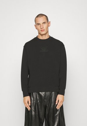 Sudadera negra de cuello redondo y mangas largas, con un sutil logotipo en relieve en el pecho. Combinada con pantalones de cuero negro brillante.