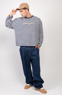 Koszula w paski z długim rękawem w kolorze granatowym i białym, z żółtą grafiką. Połączona z luźnymi niebieskimi dżinsami i brązowymi sneakersami. Beżowa czapka dopełnia całość.