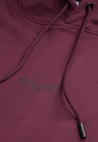 Sweat à capuche bordeaux en tissu doux, doté d'une poche kangourou à l'avant, d'une capuche avec cordon de serrage, et d'un logo brodé en noir. Texture lisse.